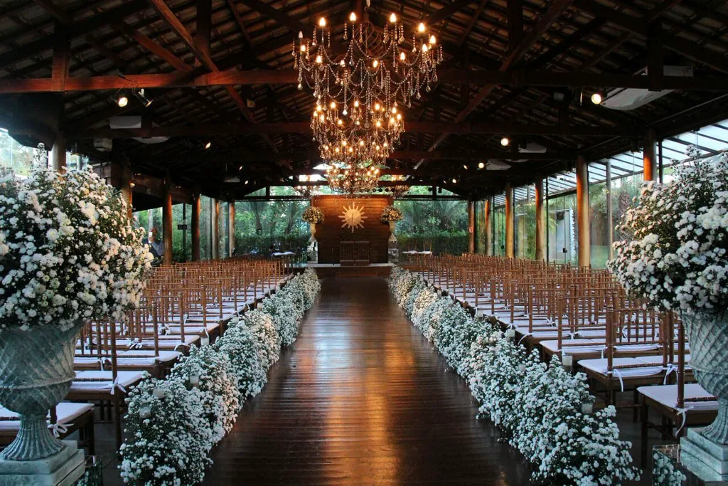 Capela da Villa do Lago com teto de madeira, lustre, cadeiras enfileiradas e corredor com flores brancas