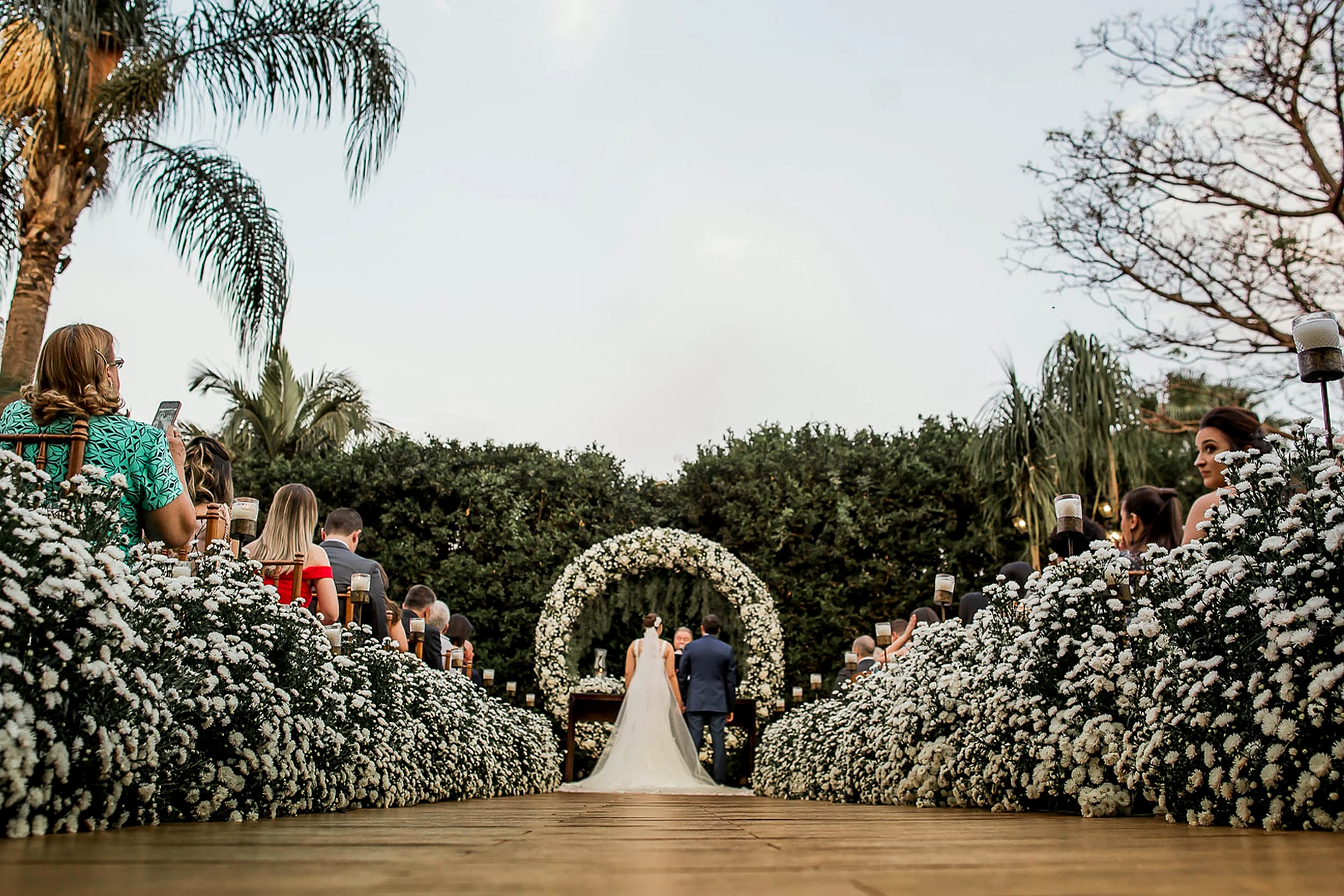 Cerimônia de casamento ao ar livre acontecendo na Villa do Lago, com corredor de flores brancas, convidados e noivos no altar