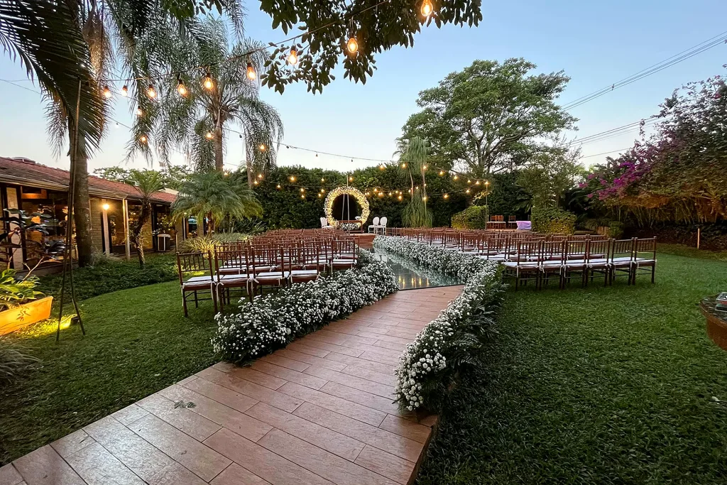 Área de cerimônia ao ar livre de dia na Villa do Lago, com gramado, palmeiras, cadeiras e arco de flores