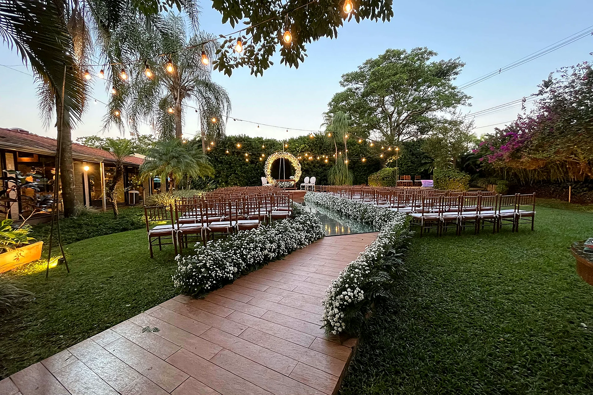 Área de cerimônia ao ar livre de dia na Villa do Lago, com gramado, palmeiras, cadeiras e arco de flores