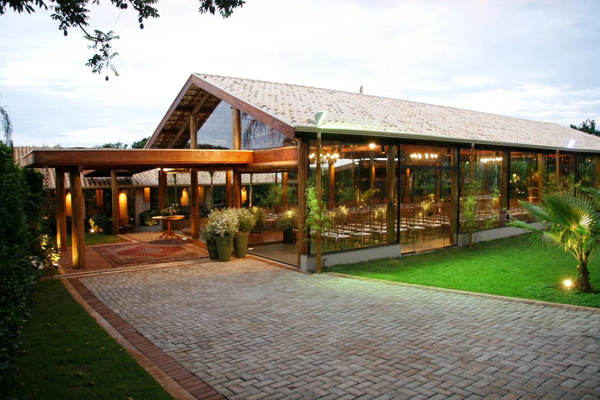 Fachada do salão principal da Villa do Lago ao entardecer, com telhado de madeira, paredes de vidro, caminho de pedra e jardim iluminado